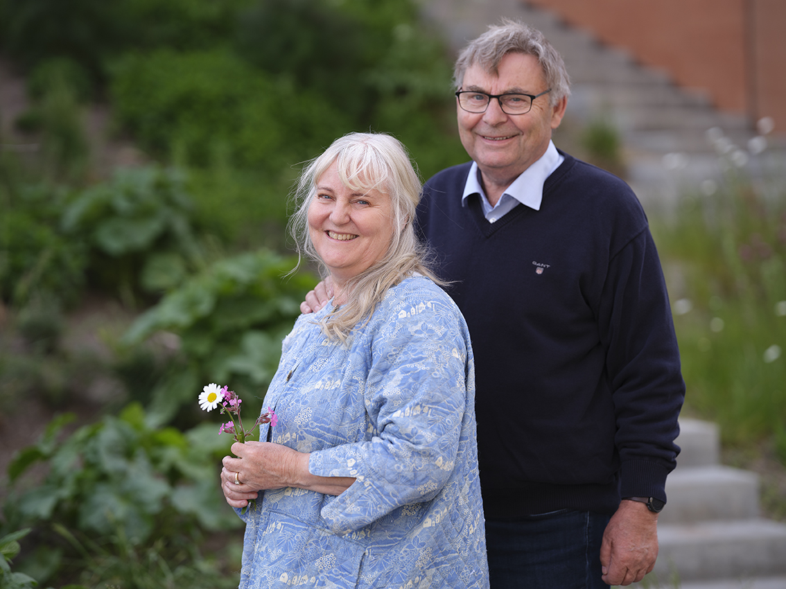Ældre ægtepar står med armene om hinanden på en sti i det grønne og kigger direkte på kameraet.