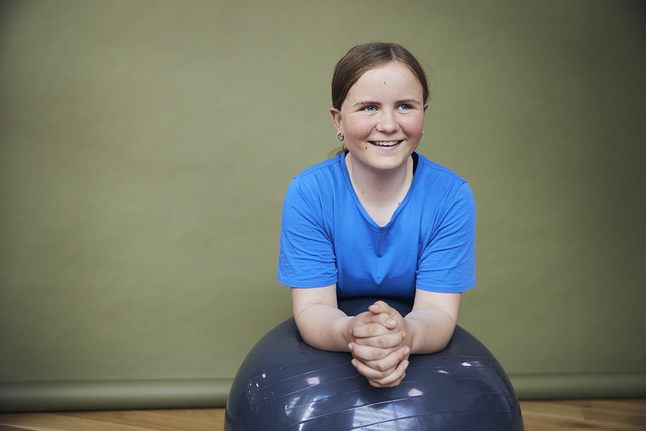 Portrætbillede af unge piger med yogabold, som hun sidder bag med hænderne hen over, mens hun smiler.