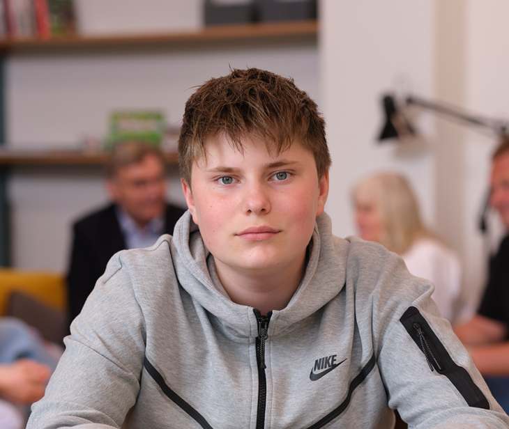 Dreng, der sidder alene, men der er menneske ri baggrunden. Han kigger direkte på kameraet.