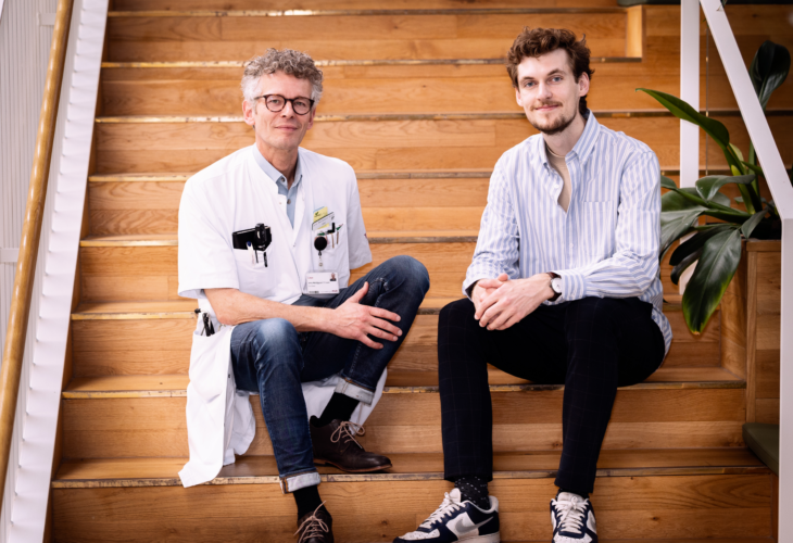 To mænd, en forsker og en læge, sidder på en trappe og kigger direkte ind i kamaraet..